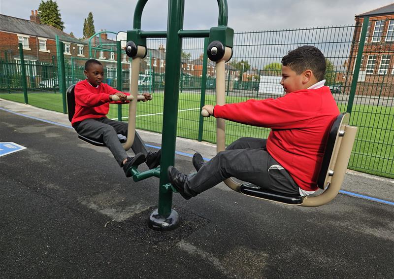 Two children are sat on a piece of gym equipment called a Seated Leg Press. The children are looking at each other and laughing as they work out.